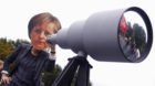 A woman wearing a mask depicting German Chancellor Angela Merkel stands behind a mock telescope during a "Freiheit Statt Angs