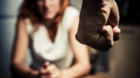 Young woman is sitting hunched at a table at home, the focus is on a man's fist in the foregound of the image